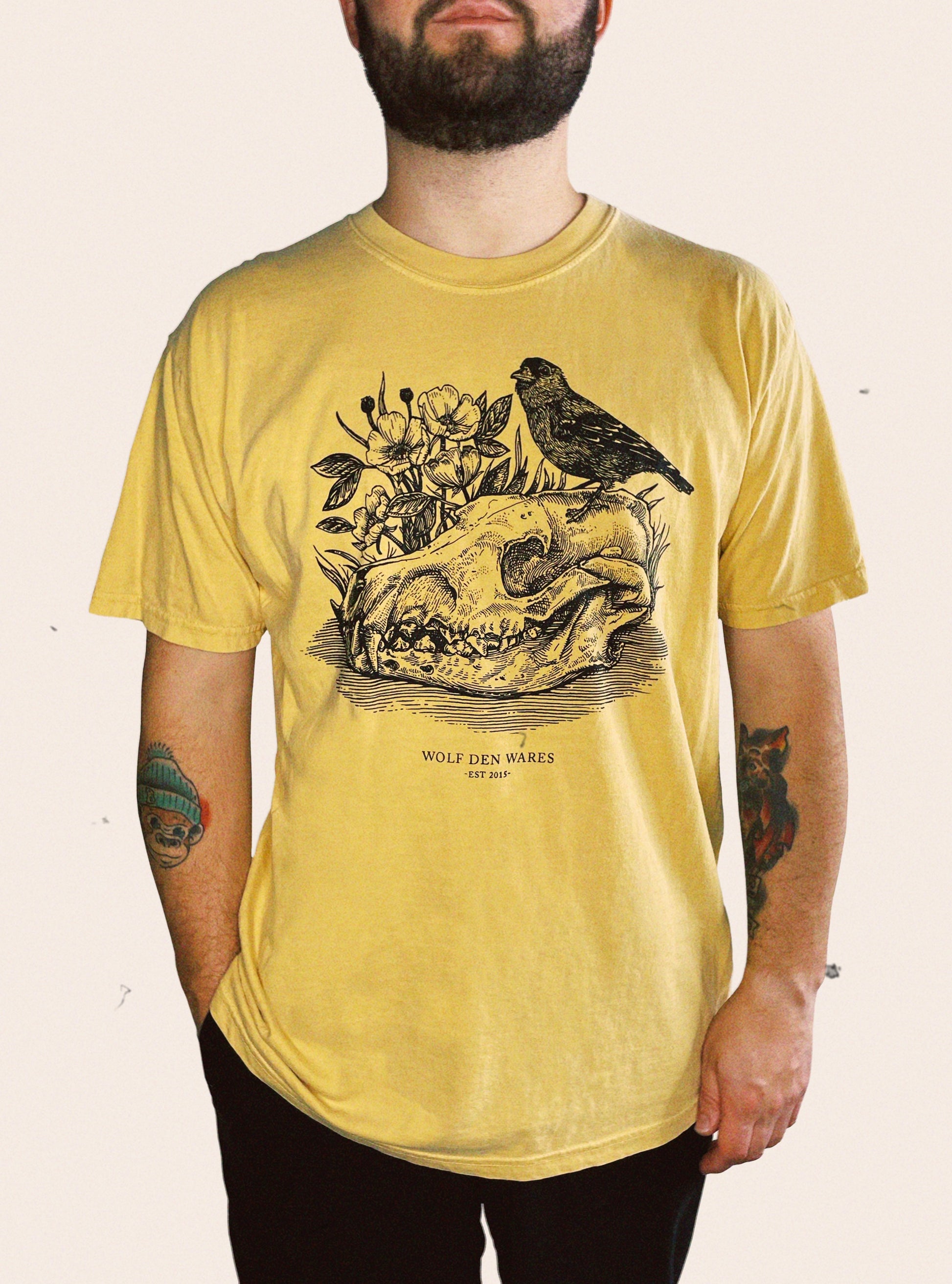 Mustard Comfort Colors tee embellished with a wolf skull, a prairie rose, and a goldfinch and Wolf Den Wares EST 2015